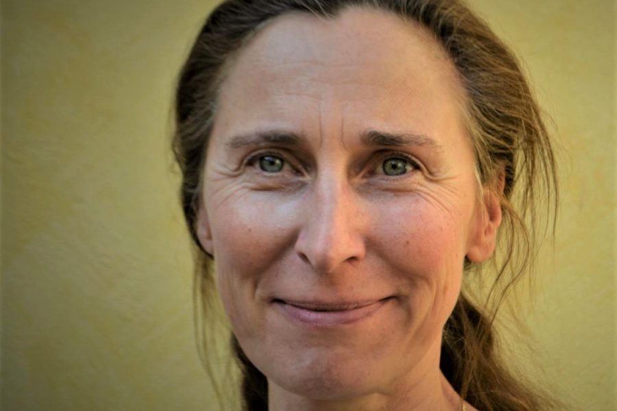 En leende kvinna med brunt hår blickar mot kameran, framför en gul bakgrund. Hennes hår är uppsatt och hon bär en blå tröja. Ljuset betonar hennes ansiktsdrag och skapar en varm atmosfär.