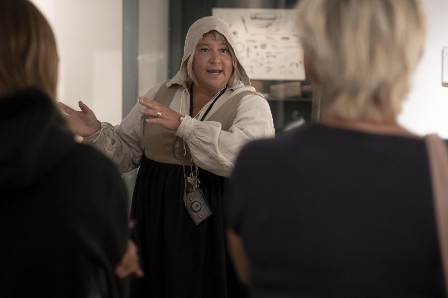 En kvinna i historisk klädsel står framför en glasmonter och talar engagerat till en grupp åhörare. Hon gestikulerar med händerna, och två personer ses bakifrån. Montern innehåller gamla artefakter i ett museum.