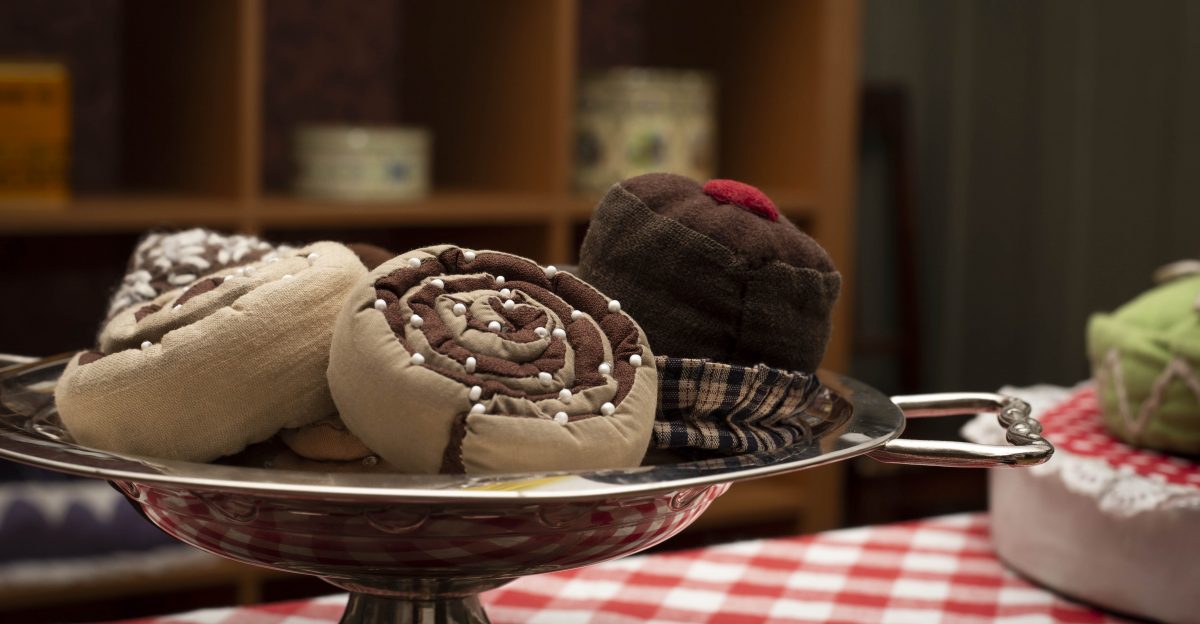 Mjuka tygkanelbullar och bakverk arrangerade på ett silverfat ovanpå en röd- och vitrutigt dukad bordsyta. Bakgrunden visar otydligt upp stora förvaringshyllor.室内のディスプレイの一部のように見えます。