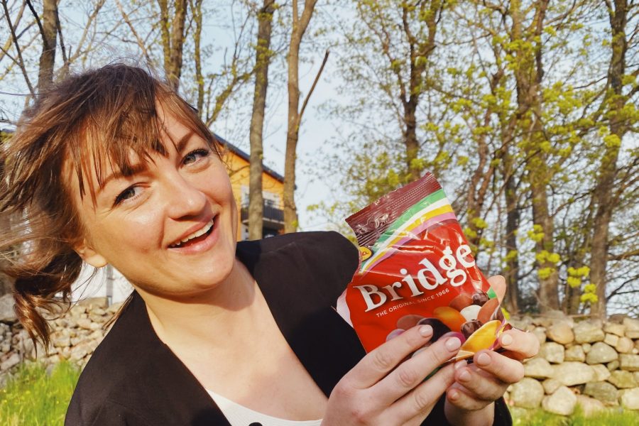 Kvinna leende och hållande en påse godis märkt "Bridge", i en utomhusmiljö med stenar och träd i bakgrunden. Förpackningen är röd med texten "The original since 1966" och visar olika godisbitar.