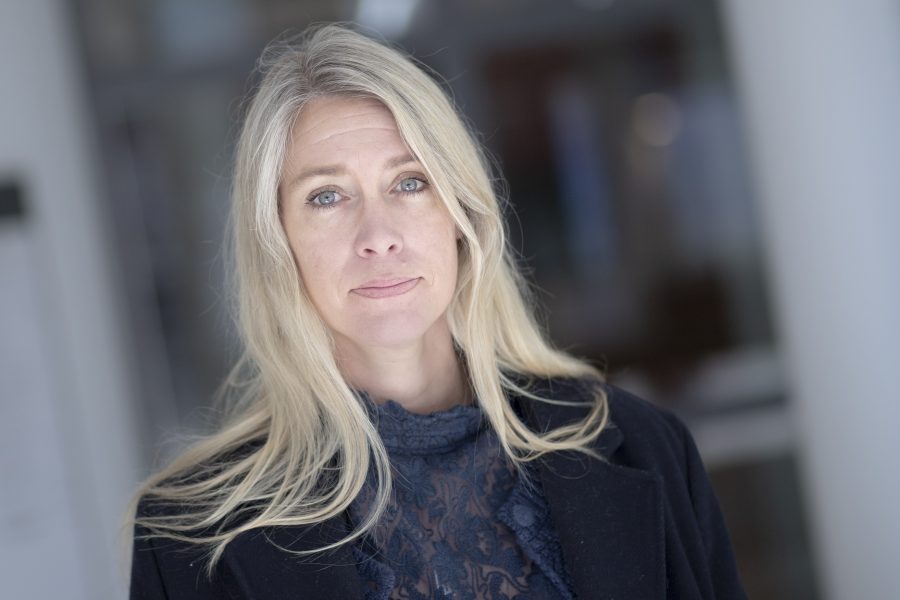 Kvinna med långt blont hår och ljus hy står och ser in i kameran, klädd i en svart kavaj och mörk spetsblus. Bakgrunden är suddig, vilket tyder på en inomhusmiljö med dämpad belysning.