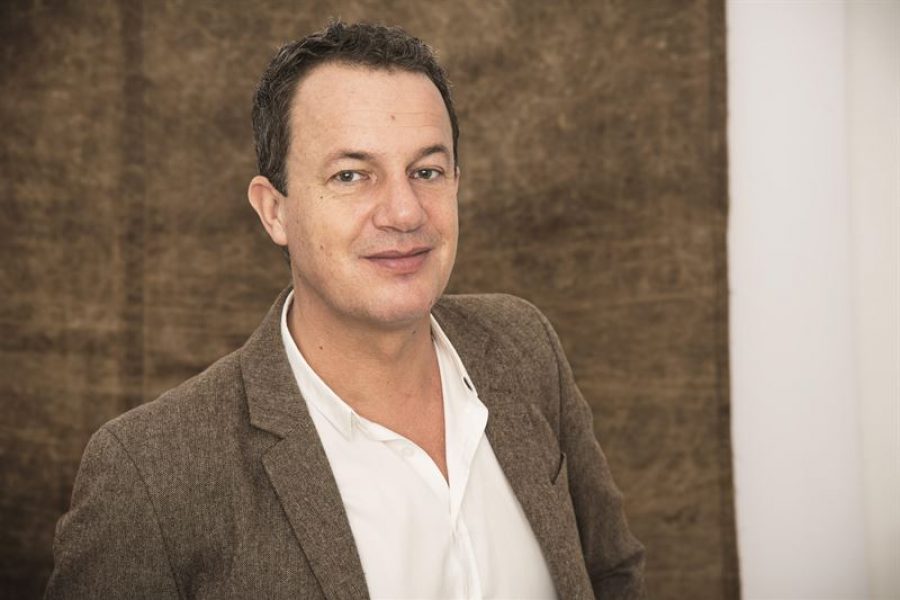 En man i en brun kavaj står framför en neutral bakgrund, tittar vänligt mot kameran. Hans vita skjorta är synlig under kavajen och han har kort, mörkt hår. Bakgrunden är oskarp och brunaktig.