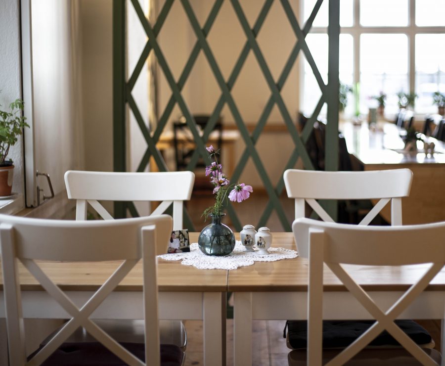 Två vita stolar vid ett träbord med en blommig spetsduk, på vilken en liten vas med rosa blommor står. I bakgrunden finns ett galler och fler bord. Ljuset strömmar in genom fönstret till vänster.