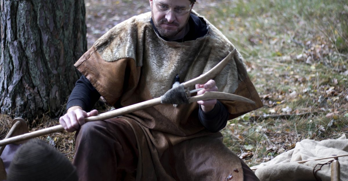 En man klädd i historiska kläder sitter utomhus i skogen vid ett träd och demonstrerar traditionellt hantverk med träverktyg för en grupp barn. Barnen sitter runt honom och är uppmärksamma på presentationen.