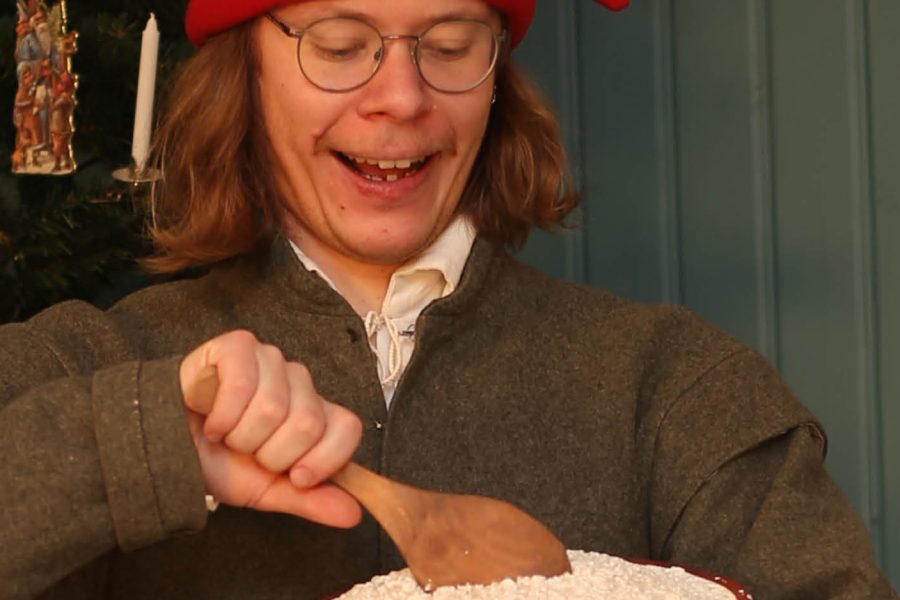 Person i röd mössa och glasögon rör i en skål med träslev, bredvid en julgran dekorerad med svenska flaggor och julpynt. I bakgrunden står en kopparkanna, och väggarna är blå. Julstämning.