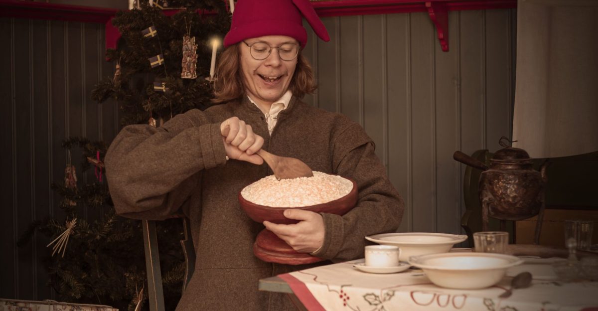 En person i traditionell svensk tomteklädsel sitter vid ett bord och rör i en skål med gröt. I bakgrunden syns en julgran och julklappar. Duken på bordet har texten "God Jul" broderad.