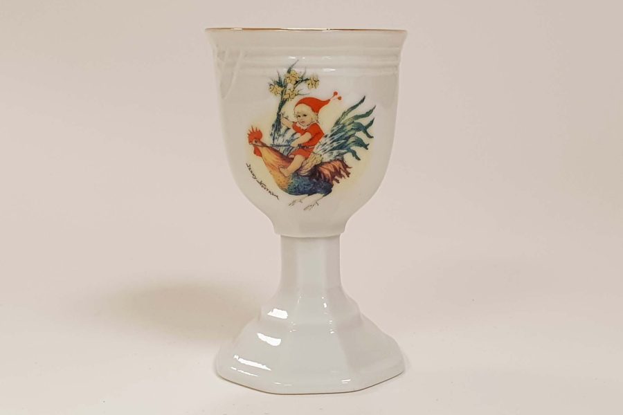 Äggkopp i porslin med motiv av en pojke i röd mössa ridande på en tupp. Designad av Jenny Nyström, med detaljer i färgerna röd, grön och brun, detaljerad mot en vit bakgrund.