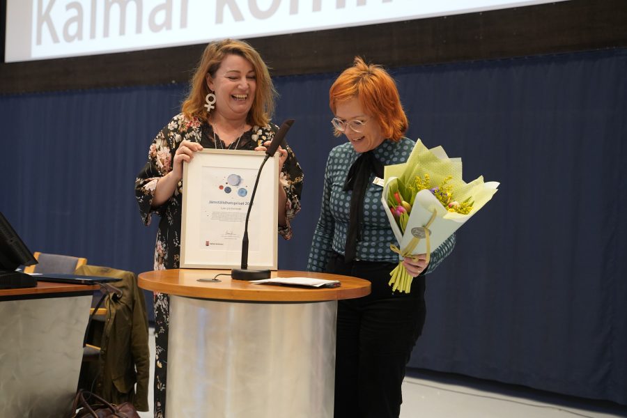 Två personer vid ett podium, en av dem håller en stor inramad utmärkelse med texten "Jämställdhetspriset 2018". Den andra personen håller en blombukett. En stolsrad och en kappa syns i bakgrunden.