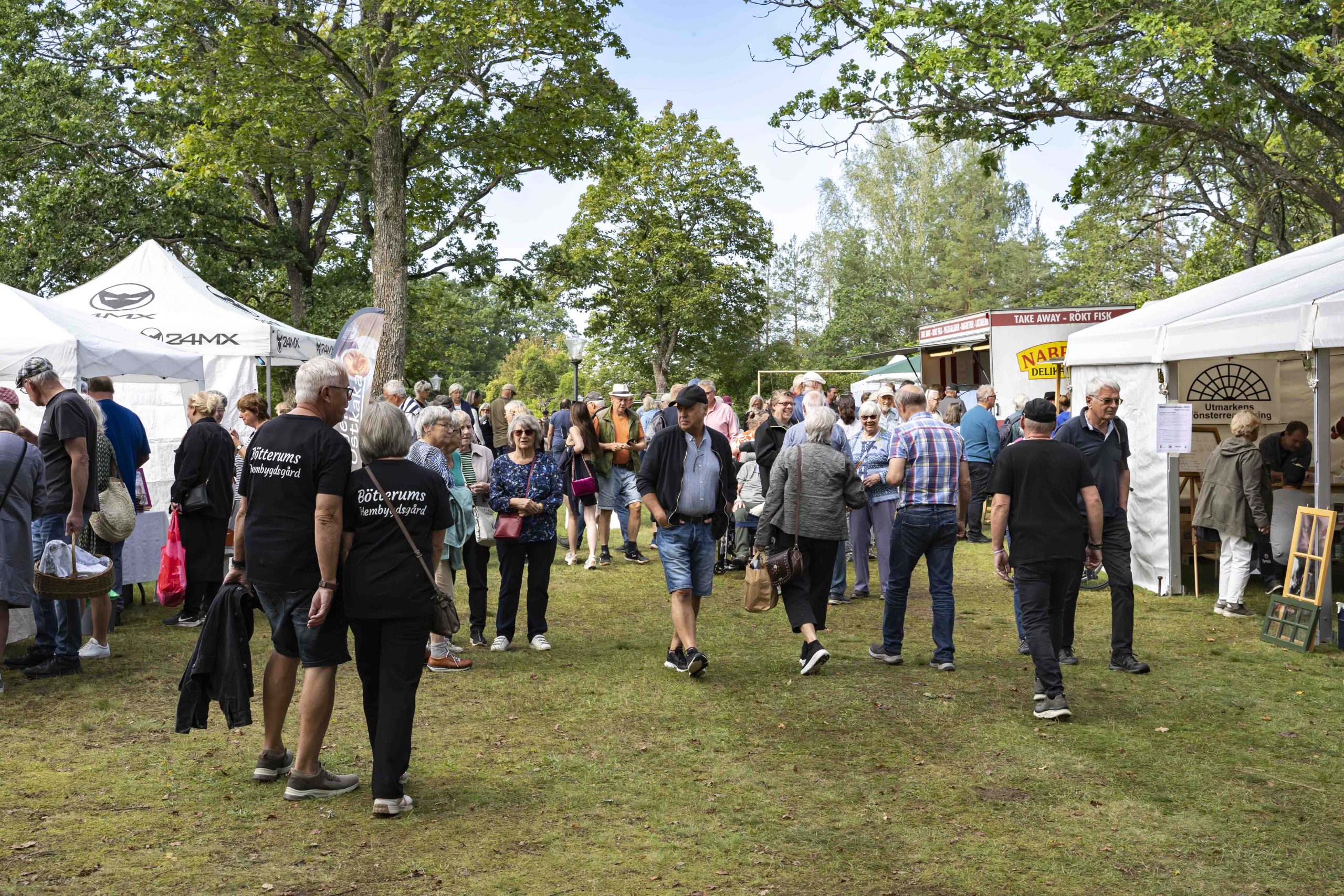 En stor folkmassa rör sig mellan vita tält under ett evenemang utomhus. Personer i svarta tröjor med texten "Börrums Hembygdsförening" minglar bland besökare. Grönskande träd omger platsen och skapar en naturlig atmosfär.