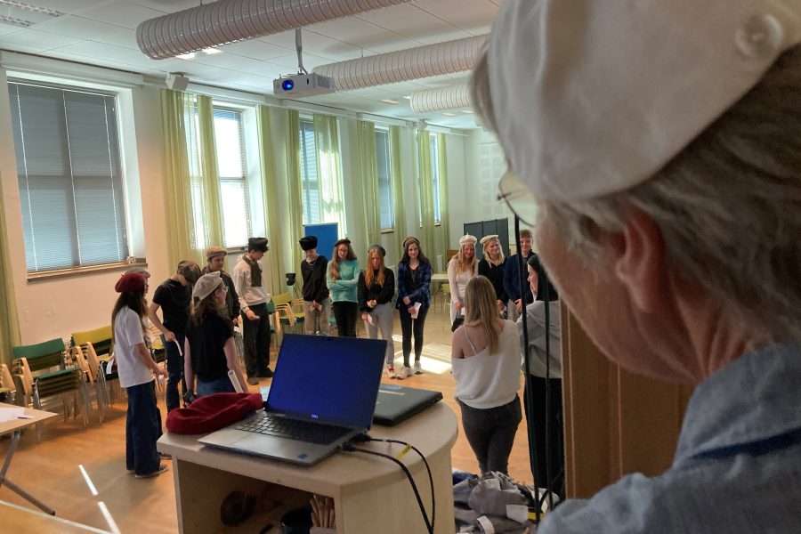 En grupp personer står i en halvcirkel i ett rum med stora fönster och gröna gardiner. De bär mössor medan en äldre person observerar från sidan nära en dator och projektor.