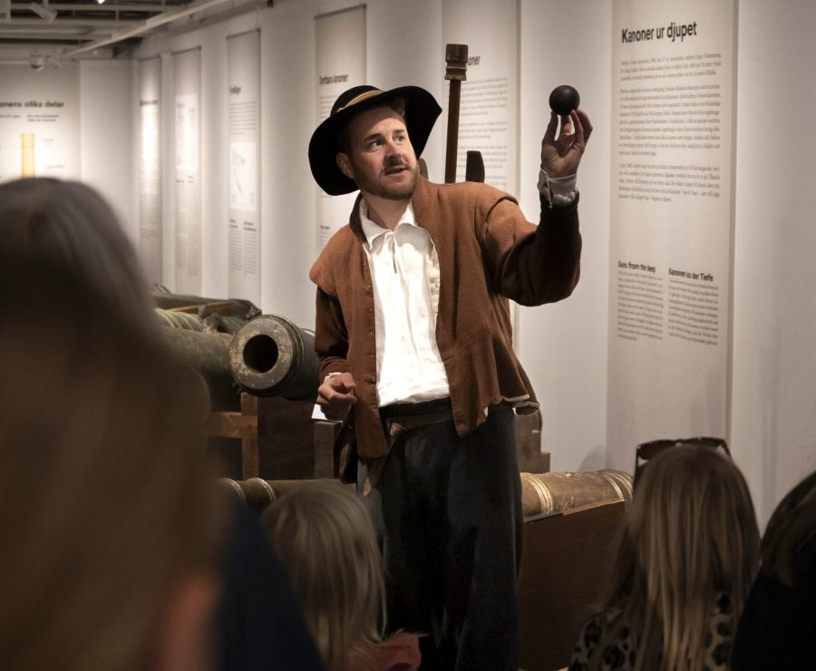 En man i historisk klädsel står i ett museum och håller upp en kanonkula inför en grupp människor, inklusive barn. Bakom honom finns utställda kanoner och paneler med texter som beskriver kanonens historia och konstruktion.