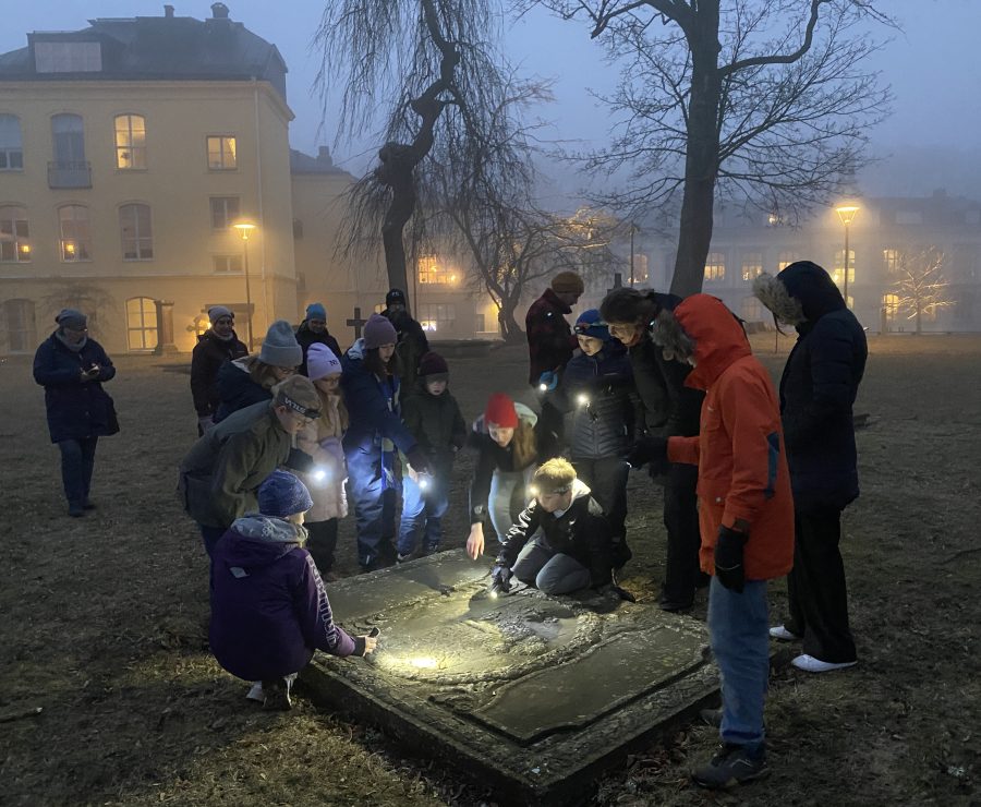 En grupp människor samlas runt en gravsten en dimmig kväll, lyser med ficklampor för att läsa inskriptionerna. Bakgrunden visar en upplyst byggnad och nakna träd, vilket framhäver den tysta och kontemplativa stämningen.
