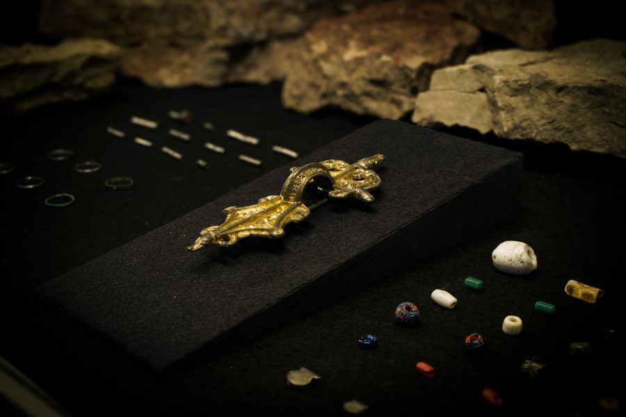 Guldfärgad spänne ligger på en svart presentationplatta omgiven av små färgglada pärlor i glas, keramik och sten. Bakom ses några oslipade klippor som bakgrund, vilket ger ett historiskt intryck och museumskänsla.