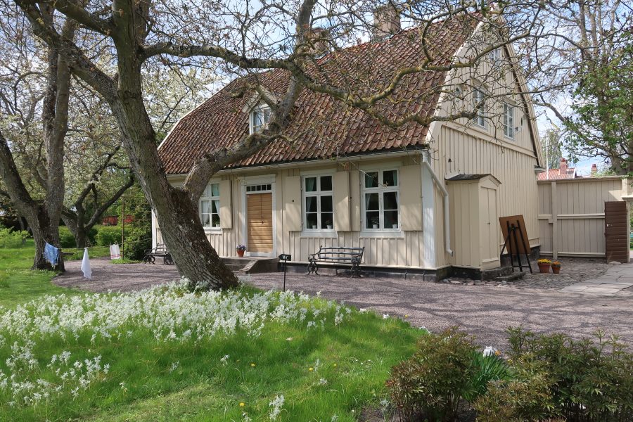 Gammalt trähus med tegeltak omgiven av blommande träd och vårblommor på gräsmattan. Tvättlinor hänger i trädgården. En grusgång leder till ytterdörren, och en skylt märkt "WC" syns bredvid huset.