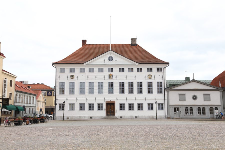 Stor vit byggnad med rött tegeltak står på ett kullerstensbelagt torg. Byggnaden har många fönster och en dekorativ klocka på fasaden. Till vänster syns ett hotell, till höger en mindre byggnad.