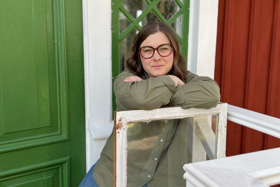 En person med glasögon lutar sig över en gammal fönsterram. Bakgrunden visar en grön dörr och en röd trävägg. Bilden kan antyda temat av fönsterrenovering, med betoning på traditionellt hantverk.