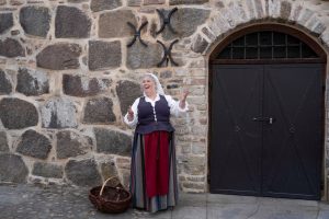 Kvinna i 1600-talsdräkt står leende mot en stenvägg bredvid en korg, framför en mörk träport i Kalmar. Tre järndekorationer pryder väggen ovanför henne, vilket förstärker den historiska atmosfären.