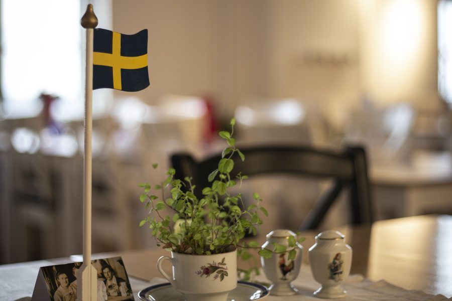 Svensk flagga på ett bord i ett kafé med gamla fotografier och ett blommande tefat. En handmålad kopp innehåller en grön växt. I bakgrunden syns suddiga konturer av stolar och bord i en ljus miljö.