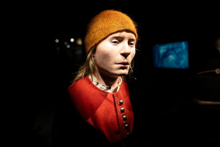 Byst av en klädd person med långt hår och orange mössa i dunkelt belyst utställning. Personen bär en röd jacka med knappar. Bakgrunden är mörk, med ett svagt upplyst blått objekt till höger.