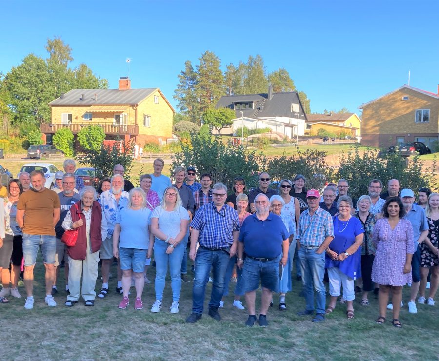 En grupp människor står utomhus på en solig dag framför några hus med träd i bakgrunden. De är klädda i sommarkläder och ser glada ut, som om de deltar i ett gemensamt evenemang.
