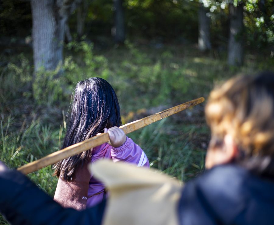 Person med långt mörkt hår och rosa jacka håller en träpinne över axeln i en skogsmiljö. En annan person med kortare hår syns delvis i förgrunden, båda vända bort från kameran. Solen skiner genom träden.