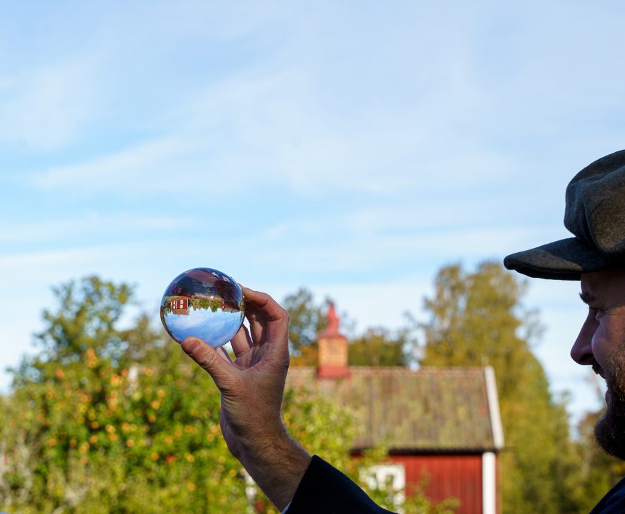 En man med keps håller en genomskinlig sfär i handen, där en spegelvänd bild av ett rött hus visas. I bakgrunden syns grönskande träd och en klar blå himmel. Solig dag på landsbygden.