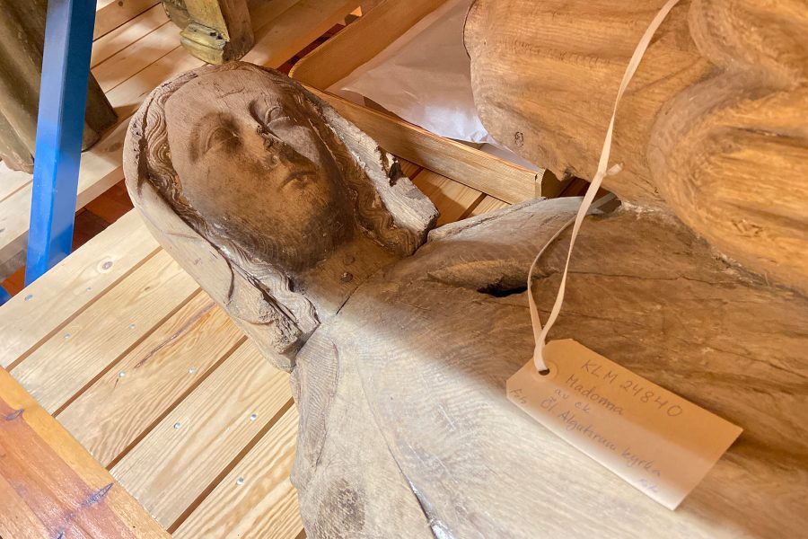 Träskulptur föreställande en liggande Madonna, placerad på ett träbord. Skulpturen har detaljerade ansiktsdrag och en lång klädedräkt. En etikett med text: "KLM 24840 Madonna av ek från gamla Algutsrum kyrka" är fäst vid henne.