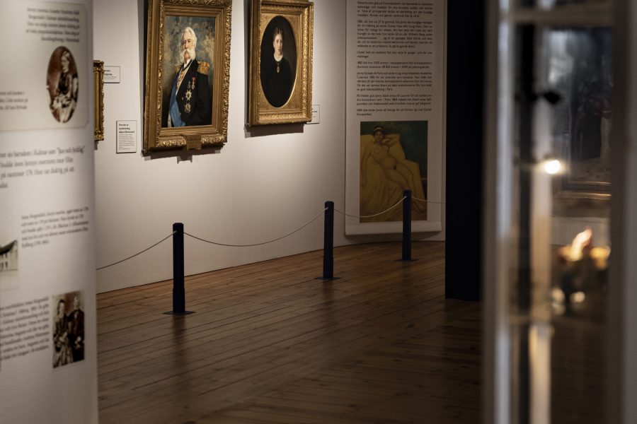 Konstgalleri interiör med tavlor i guldramar, upphängda på en vit vägg. Trägolv reflekterar belysning, och glasmonter med små föremål syns delvis till höger. Informationspaneler erbjuder historisk kontext på vänster sida.