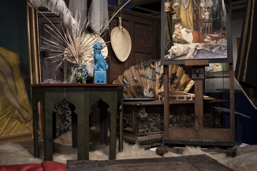 Ett konstnärligt arrangemang i ett rum med ett staffli som visar en målning av en liggande person och en vit hund. I förgrunden finns orientaliska dekorationsobjekt, inklusive en blå staty, fjädrar och fläktar.