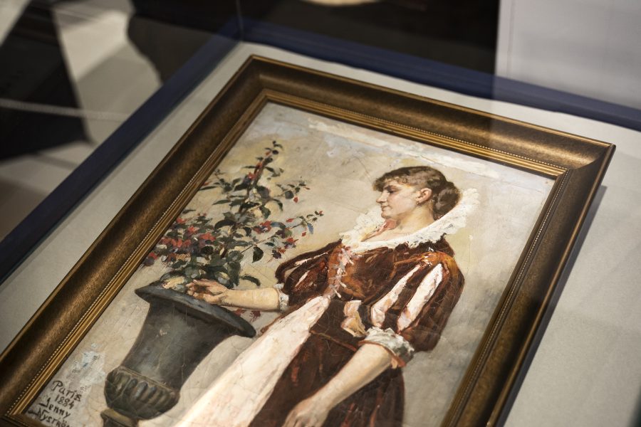 Målning i glasmonter visar kvinna i röd klänning med ett blomsterarrangemang i dekorativ vas. Tavla signerad Paris 1887, Jenny Nystrom. I bakgrunden, färgglatt utsmyckad solfjäder. Utställning i museimiljö.