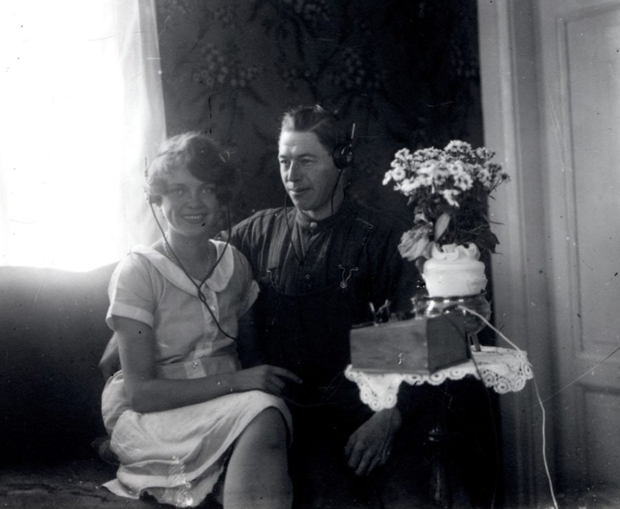 En man och en kvinna sitter tillsammans i ett rum, båda bär hörlurar. Kvinnan ler, medan mannen ser koncentrerad ut. Bredvid dem står ett bord med en vas med blommor och en radioapparat.
