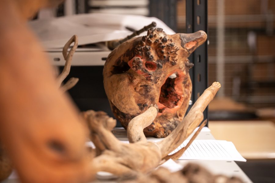 Förvridet trä och bark med komplexa texturer och håligheter ligger på en metallhylla. Omfattande brun och röd färg markerar ytan. Ospecifika papper syns i bakgrunden. Miljön verkar vara ett lagerutrymme med diverse föremål.