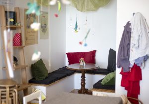 Hörnsoffa med svarta dynor och färgglada kuddar omger ett runt bord av trä. Väggarna pryds av affischer. Möbler och kläder hänger intill. Flera mobiler dinglar ovanför med olika färger och former.