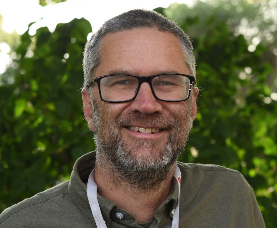 En man med kort grått hår och skägg ler in i kameran. Han bär svarta glasögon och en grön skjorta. I bakgrunden syns gröna löv och suddigt solljus skiner igenom bladen.