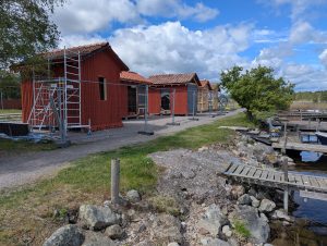 Röda träbodar under renovering står på rad längs en grusväg vid en sjö. Byggnadsställningar är uppställda bredvid en bod, och en träbrygga leder ner till vattnet. En grönskande buske skymtar i bakgrunden.