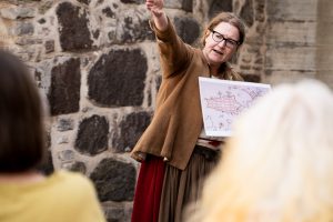 En kvinna i historisk dräkt står framför en stenmur och gestikulerar medan hon håller ett kartblad. Två personer i förgrunden lyssnar uppmärksamt. Scenen antyder en guidad tur eller berättelse utomhus.