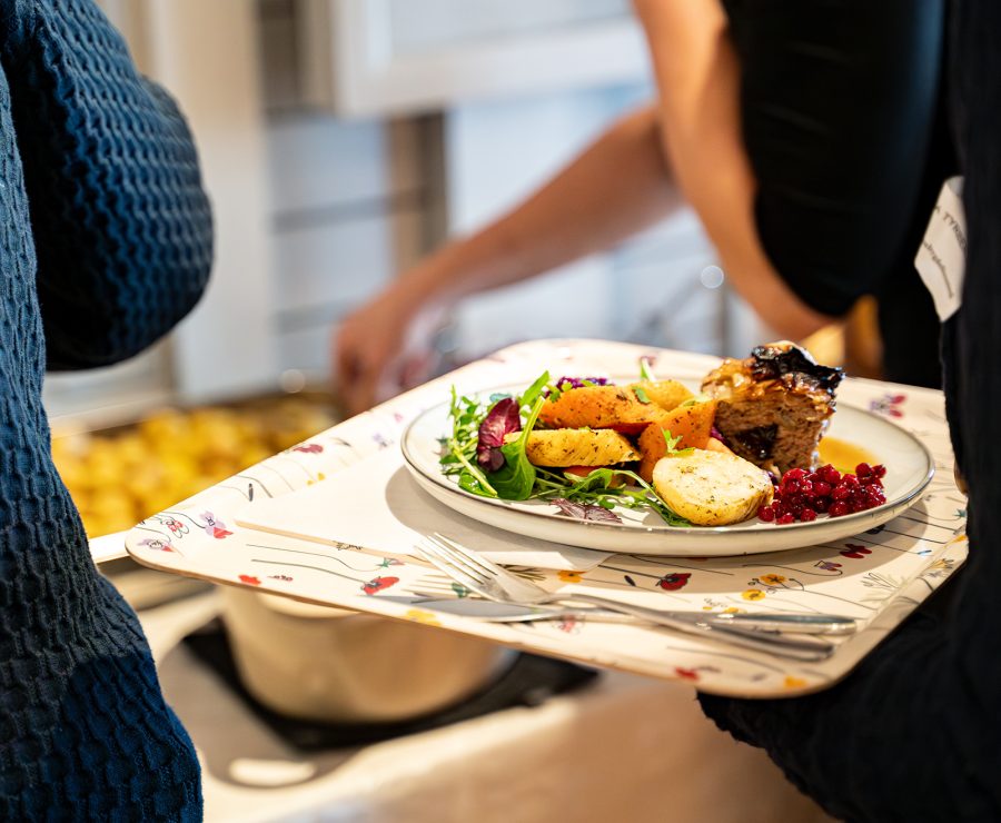 En person bär en bricka med en tallrik innehållande rostad potatis, kött, sallad och lingon. En annan person serverar i bakgrunden, inomhusmiljö. Brickan är dekorerad med blommönster och bestick syns bredvid tallriken.