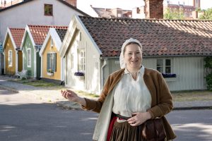 Kvinna klädd i historisk dräkt står leende framför en rad färgglada trähus med tegeltak. Solen skiner, och blomlådor med blommor pryder husen. I bakgrunden syns äldre byggnader.