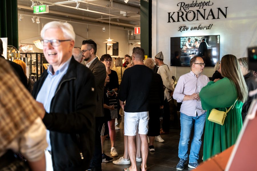 En livlig grupp människor samtalar i ett museum eller utställning, där en skylt med texten "Regalskeppet Kronan - Kliv ombord!" syns på väggen. En skärm visar en historisk filmsekvens.