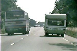 Buss och husvagn kör på en landsväg kantad av träd och grönska under en klar himmel. I bakgrunden syns flera bilar på avstånd. Bussens text är delvis läsbar och landskapet är sommargrönt.