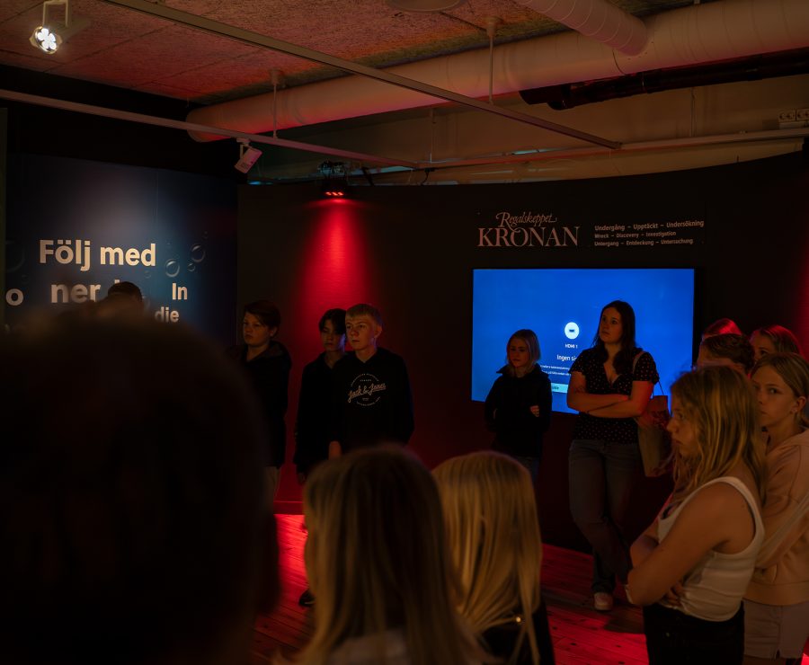 En grupp ungdomar står i ett mörkt utställningsrum med skylten "Regalskeppet KRONAN" och texten "Följ med" på väggen. Rummet är belyst av röda ljus och en skärm visar text. De uppmärksammar något utanför bilden.