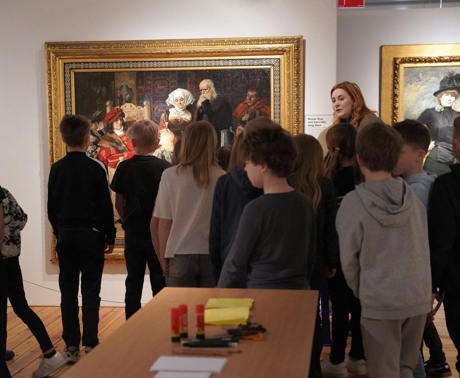 En grupp barn står framför en tavla i ett konstgalleri, guidade av en vuxen kvinna. Tavlan visar en historisk scen i en gyllene ram. Bordet i förgrunden är fyllt med färgpennor och papper.