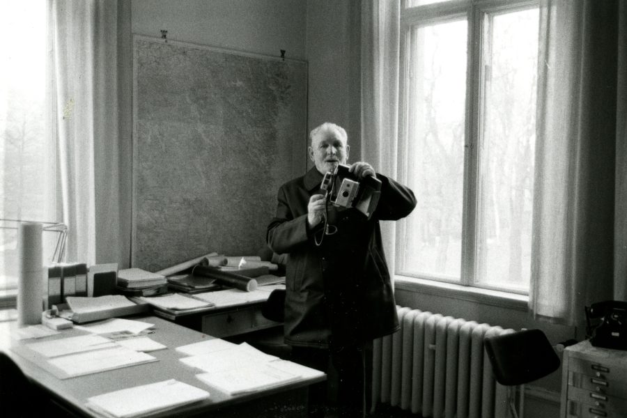 En äldre man i en kavaj håller en gammaldags kamera framför sig i ett kontorsrum med ett stort fönster som släpper in ljus. På skrivbordet ligger papper, mappar och rullar, vilket ger en känsla av pågående arbete.